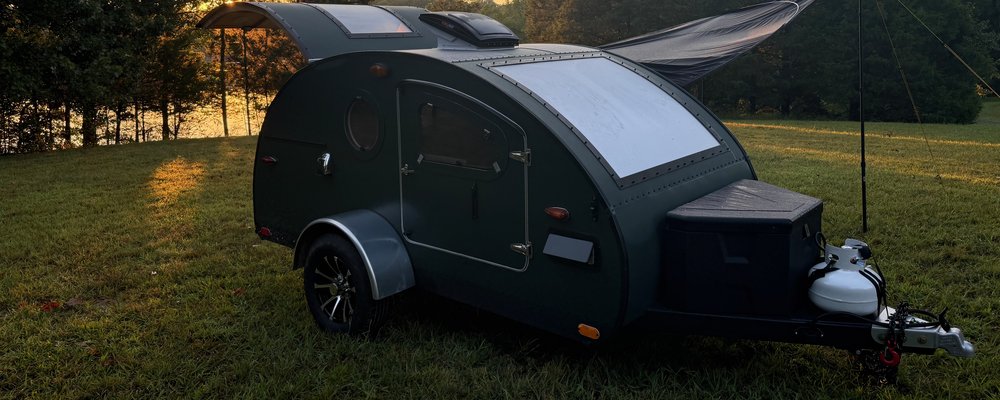 A green teardrop camper at dusk in a lakeside field, galley hatch raised, the sun setting through the trees behind it