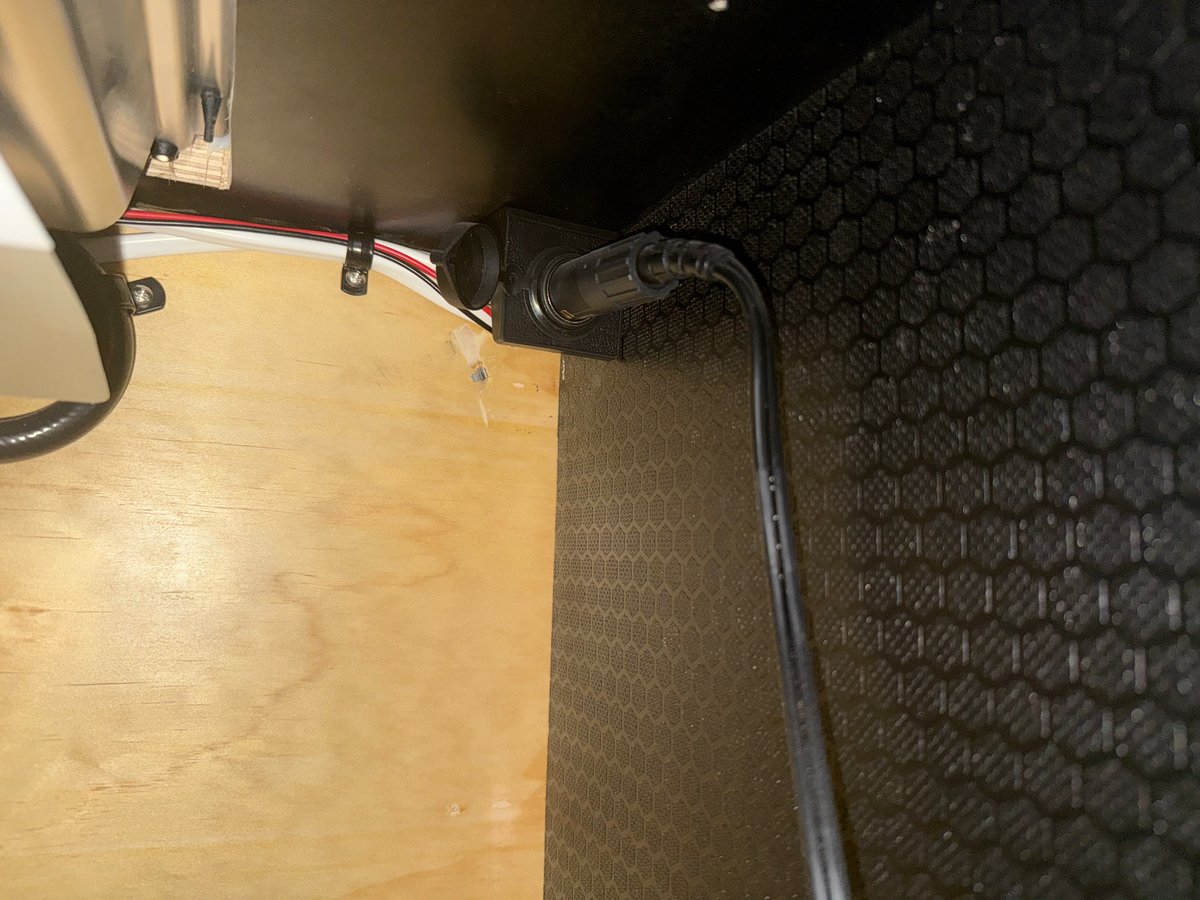 A 12V socket mounted high on the back wall of a teardrop galley bay, the fridge cable plugged in and dropping down, a red and white wire run clipped along the wall above