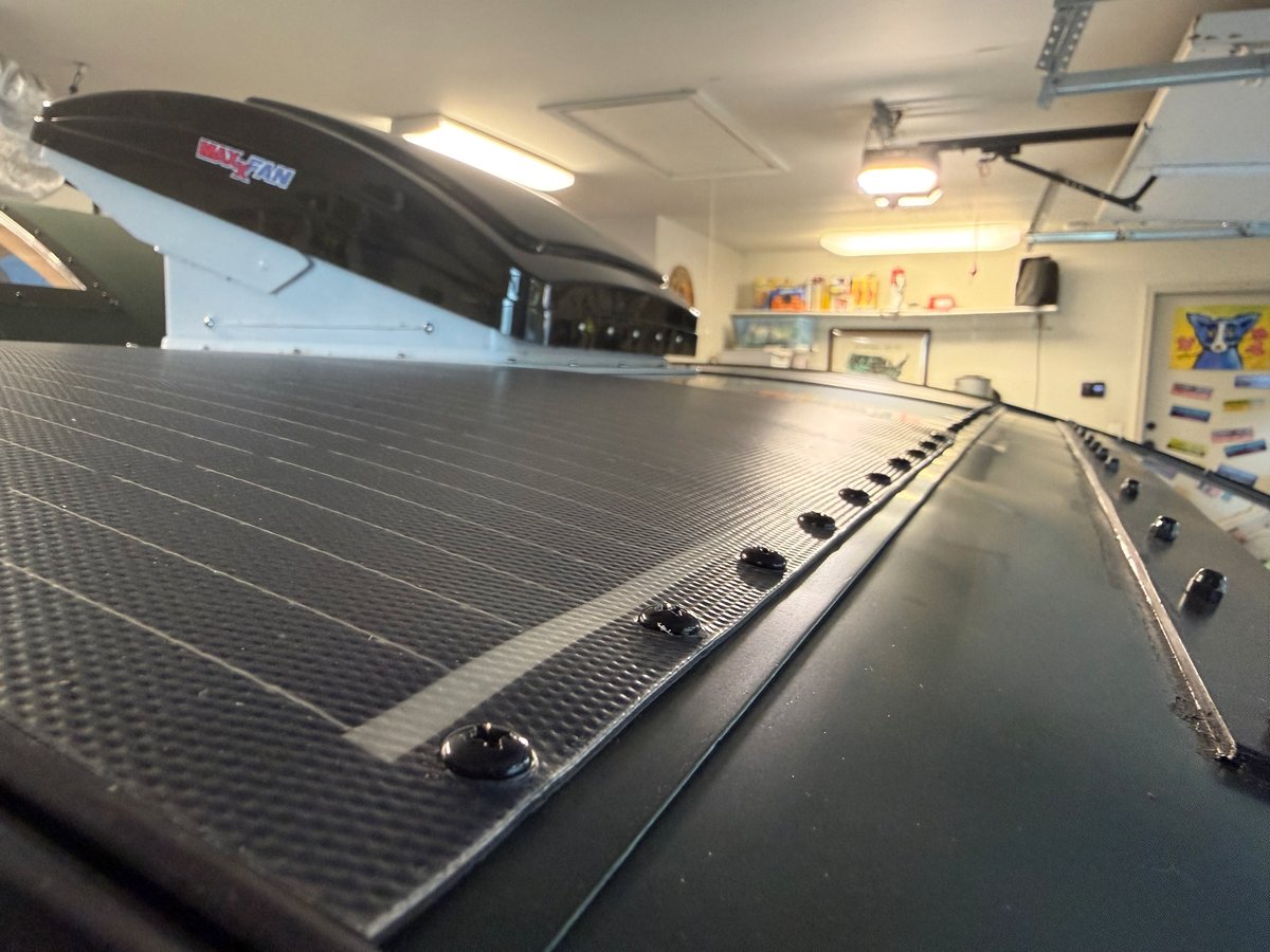 A row of black button-head screws along the front edge of a flexible solar panel on a teardrop roof, with a MaxxFan behind