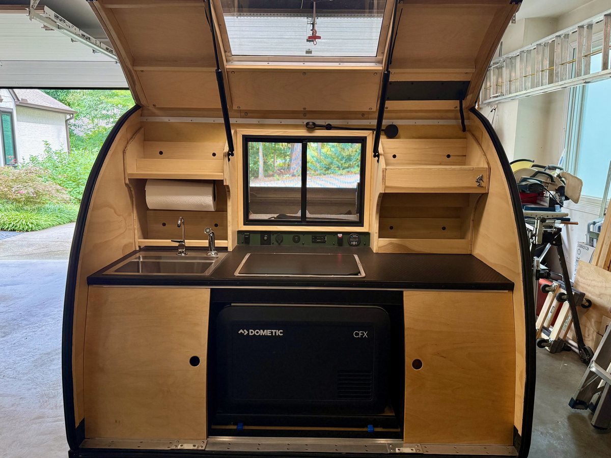 A teardrop galley with the hatch up: sink on the left, cooktop and control panel in the middle, the fridge in its bay below, drawers on the right