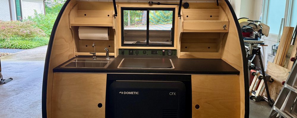 A teardrop camper galley with the hatch raised, showing the sink, cooktop, control panel and fridge bay across the full width of the counter