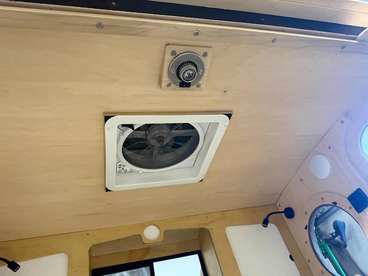 Looking up at the ceiling of a birch teardrop cabin at a white roof fan housing, with a reading light and a porthole window nearby