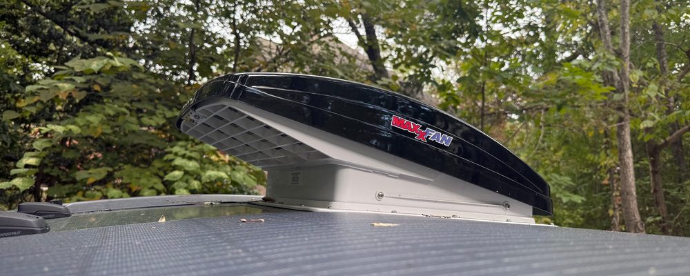 A black MaxxFan roof vent with its lid propped open on a teardrop roof, trees behind it