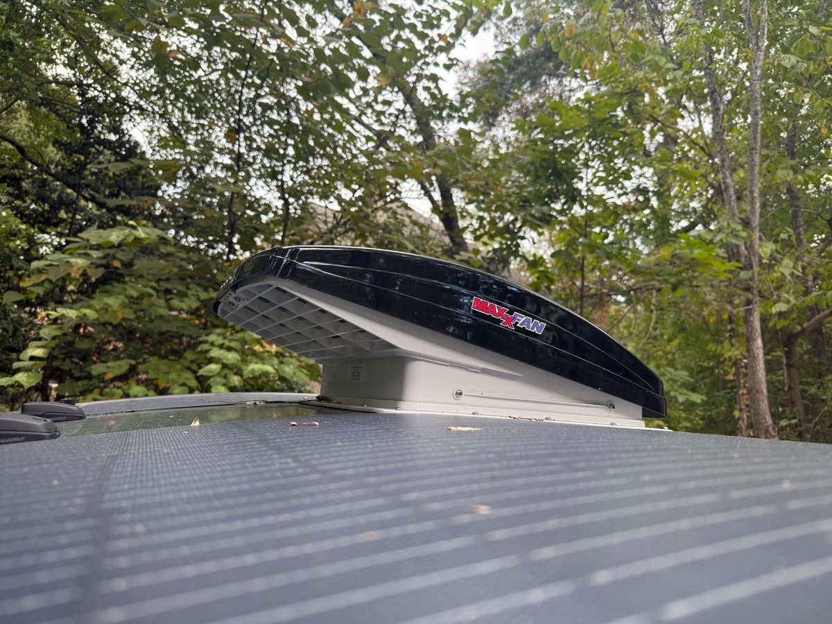 A black MaxxFan lid propped open on a teardrop roof next to a flexible solar panel, with trees behind
