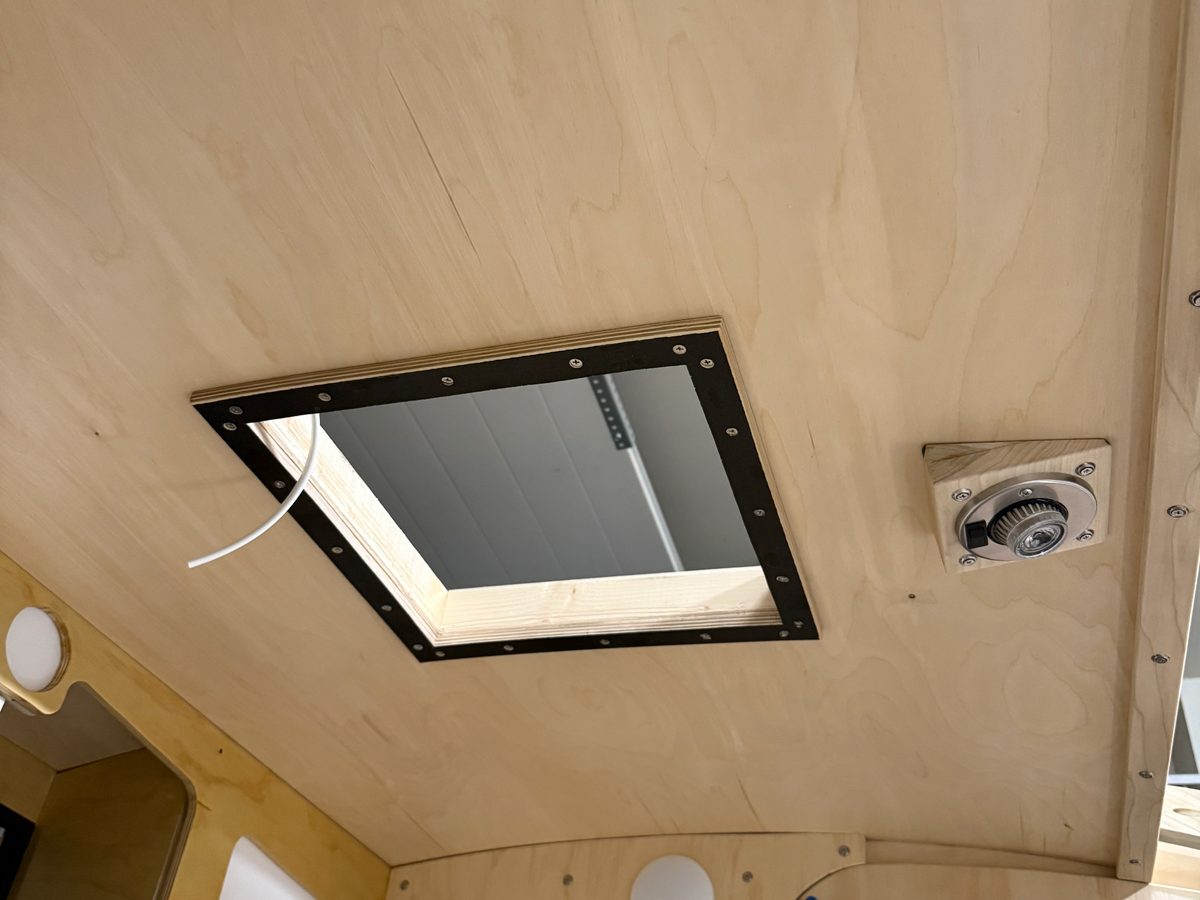 The square roof fan opening framed in black, seen from inside an unfinished birch teardrop cabin, with a wire run beside it