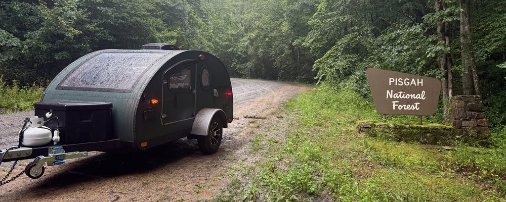 A green teardrop trailer parked on a wet gravel forest road beside the Pisgah National Forest sign