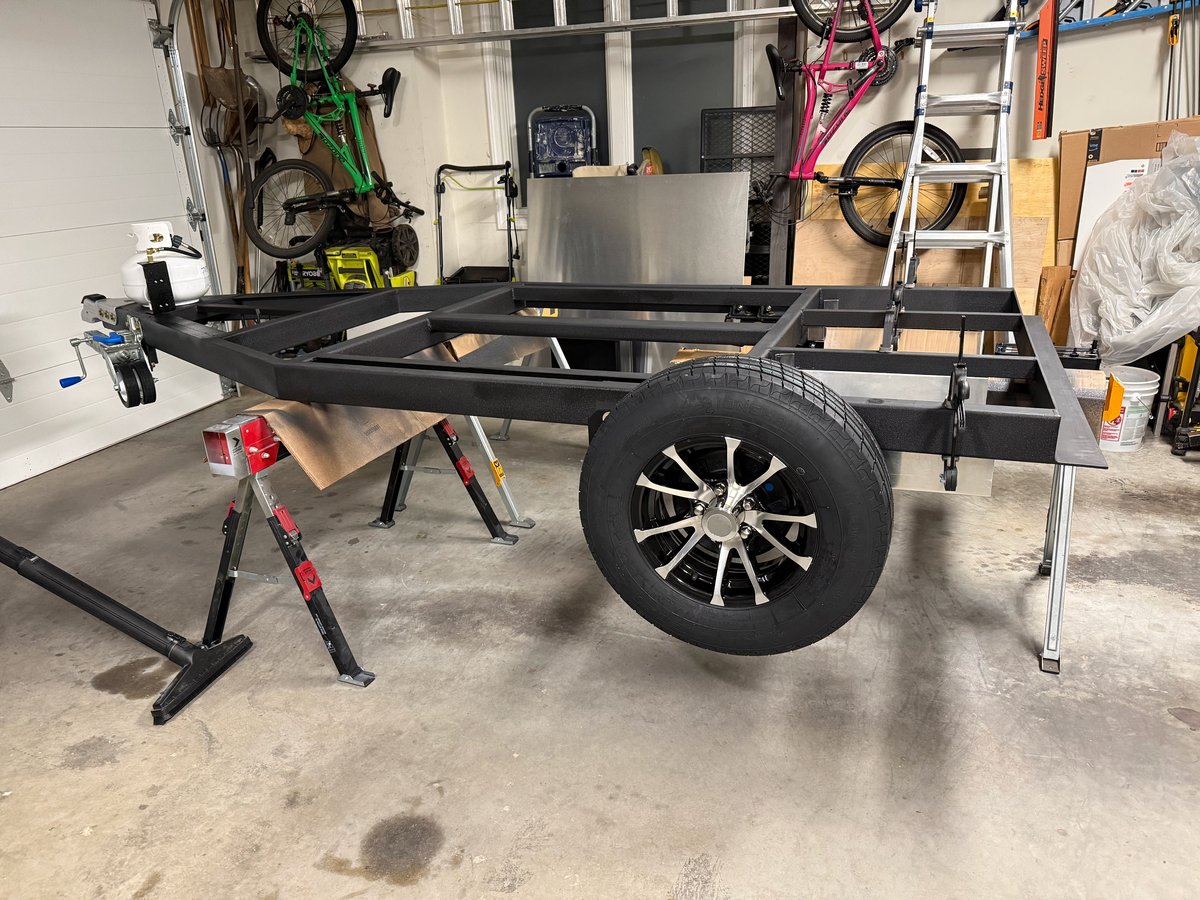 A black steel teardrop trailer frame on stands in a garage, seen from the side, with a wheel and tire mounted and open space under the frame where an axle would be