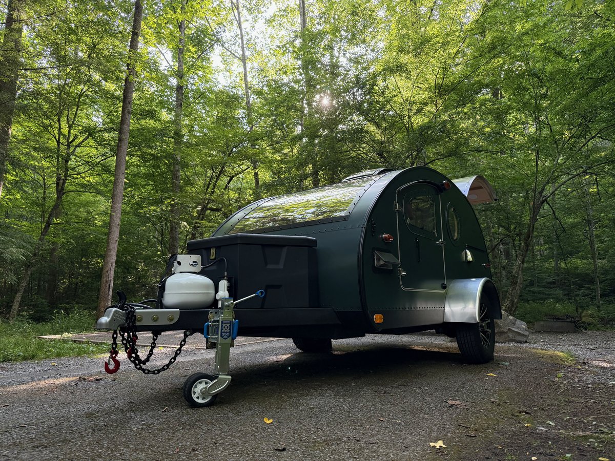 A green teardrop trailer hitched up and parked on a gravel road in thick green woods