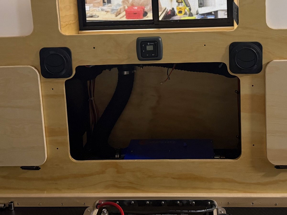 Two round heater outlets on either side of a Propex digital thermostat on a teardrop's plywood bulkhead, above an open bay showing the ducting and the heater