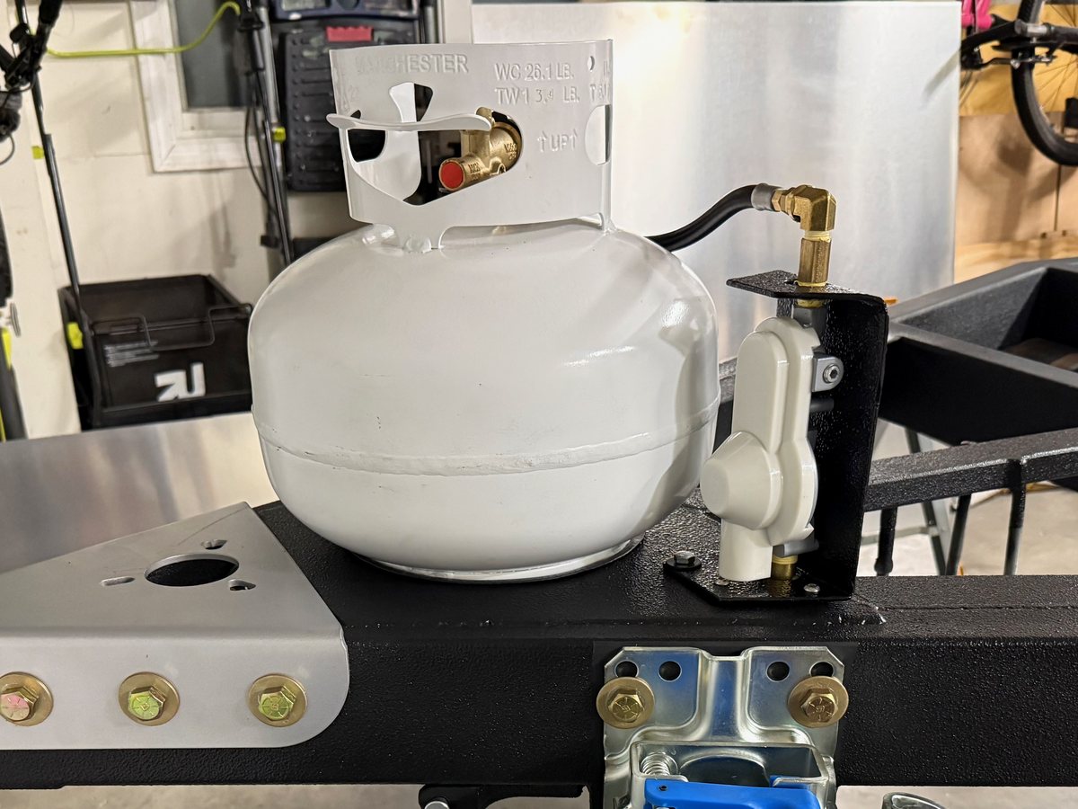 A white 11 lb squat propane tank mounted on a teardrop trailer's tongue, with a two-stage regulator bolted to a bracket beside it
