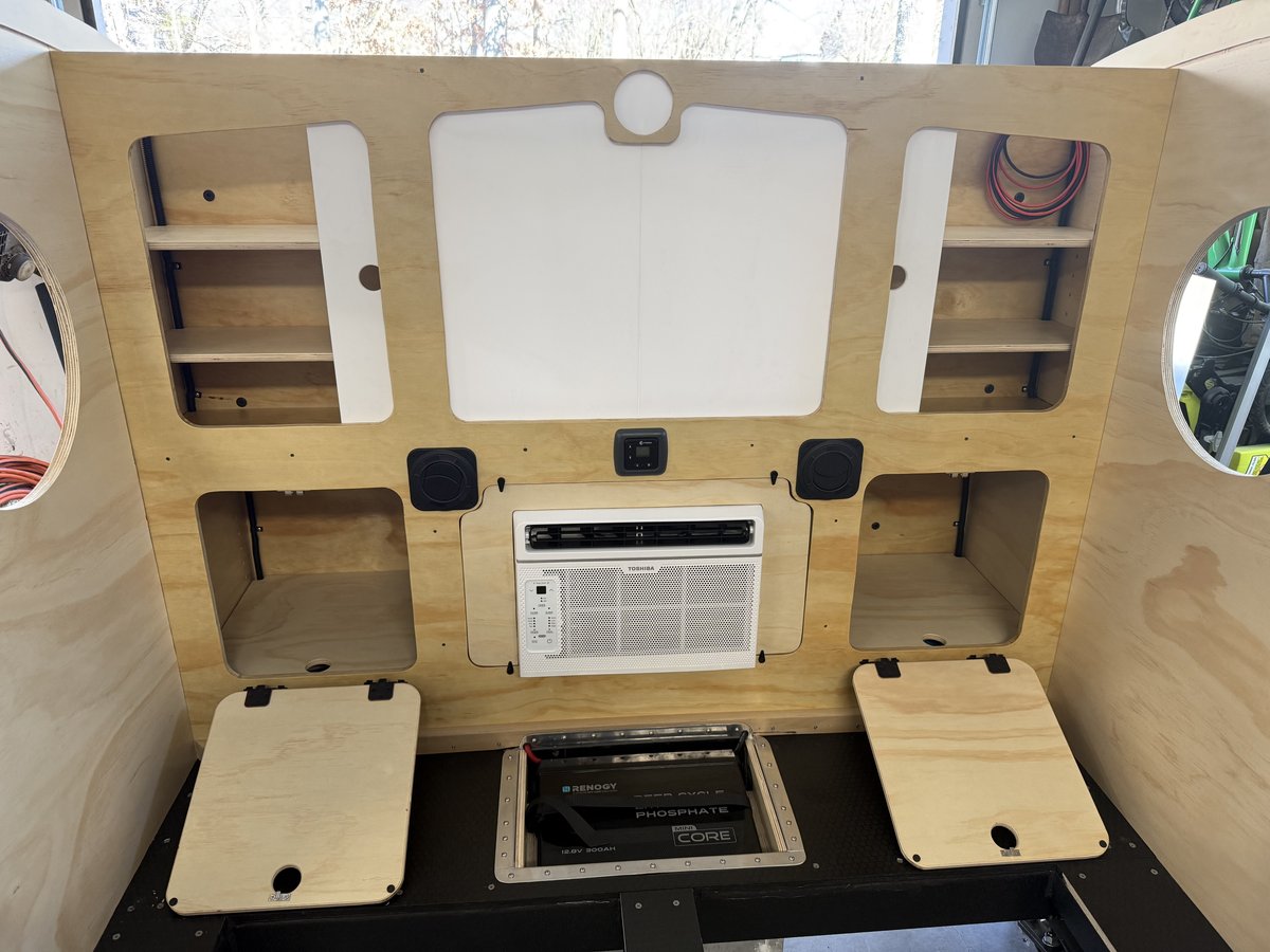 A white Toshiba window air conditioner mounted in the middle of a plywood cabinet wall inside a teardrop, with a lithium battery visible through a floor hatch below