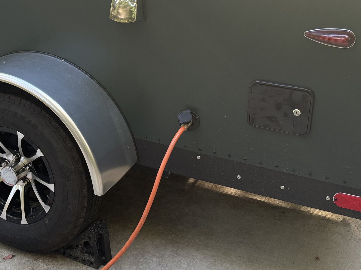 An orange extension cord plugged into a round black shore power inlet on the green side of a teardrop, just behind the fender