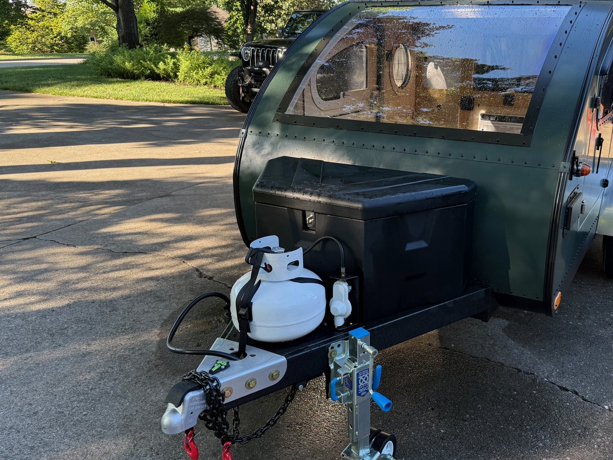 The tongue of a green teardrop camper carrying a propane bottle, a black storage box and the jack, parked on a driveway