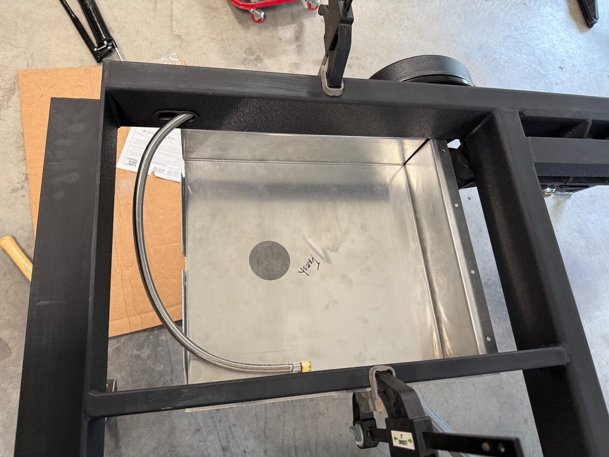 An empty aluminum pan marked Fresh set into a black steel teardrop trailer frame beside the wheel, a braided water line run through the frame