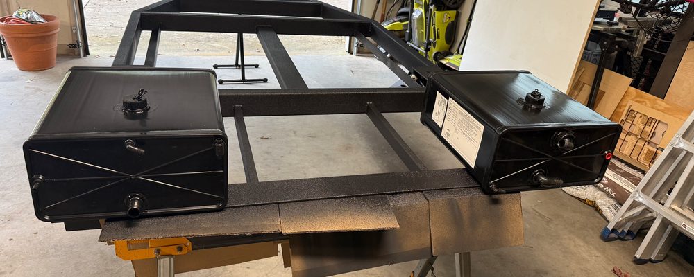 Two black 9 gallon water tanks set on a black steel teardrop trailer frame in a garage, before they went in under the galley end