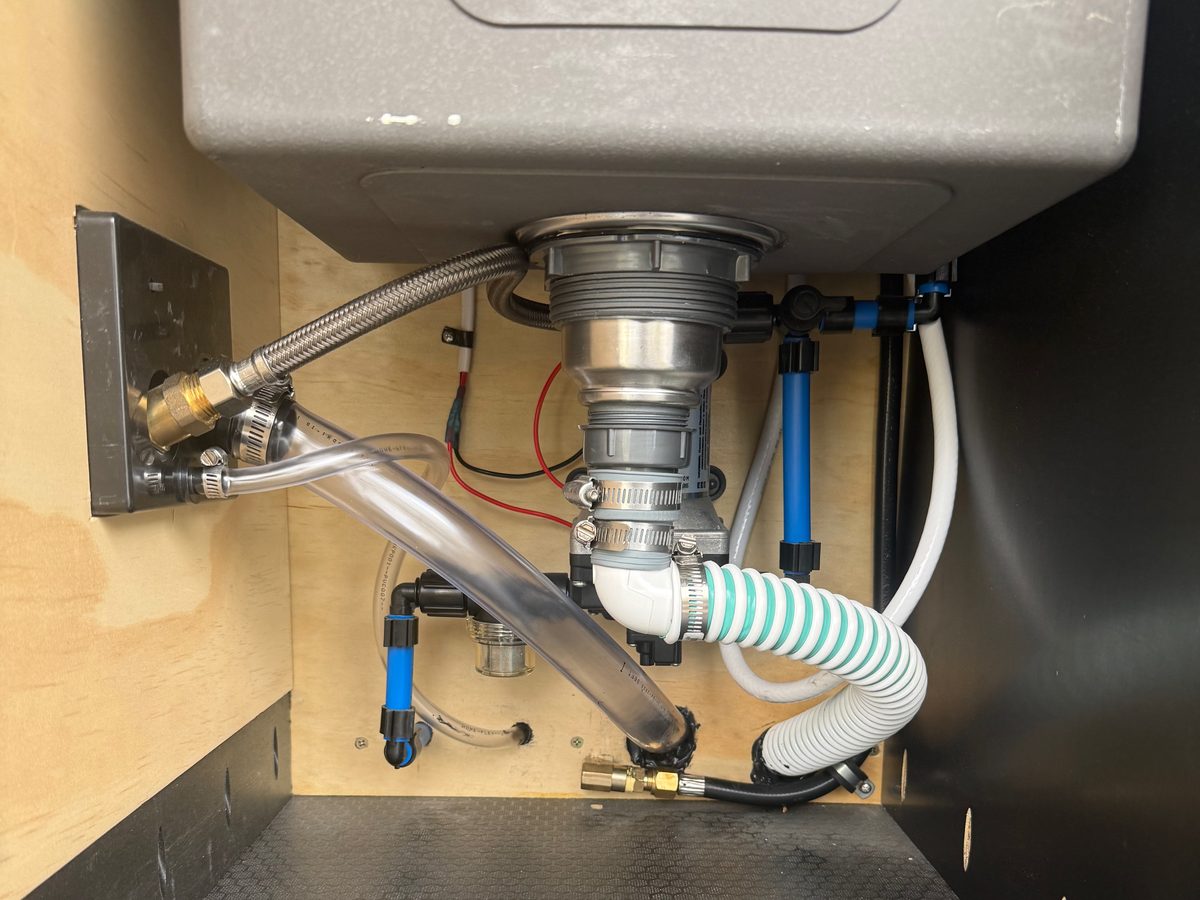 Looking up into the cabinet under a teardrop galley sink at the drain, water lines and fittings, with a black hex-pattern floor