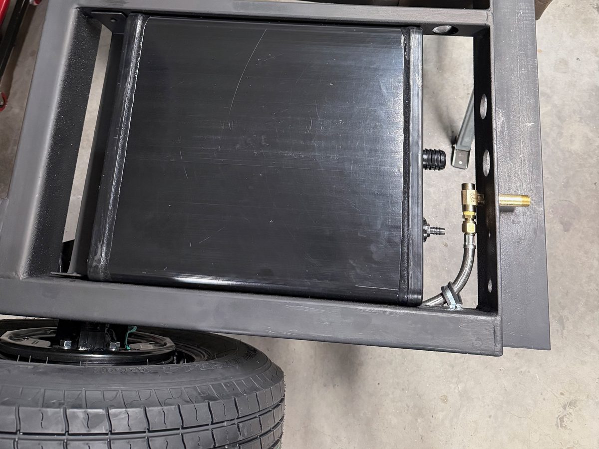 A black 9 gallon water tank seated in its pan in a black steel trailer frame, the trailer tire just below it