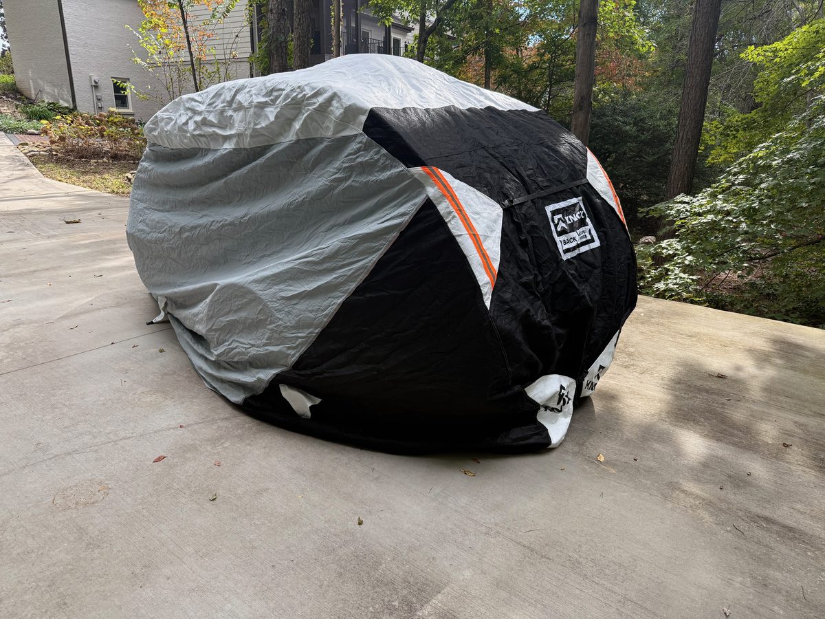 A teardrop trailer under a fitted gray and black cover with orange reflective strips, parked on a concrete pad with trees behind