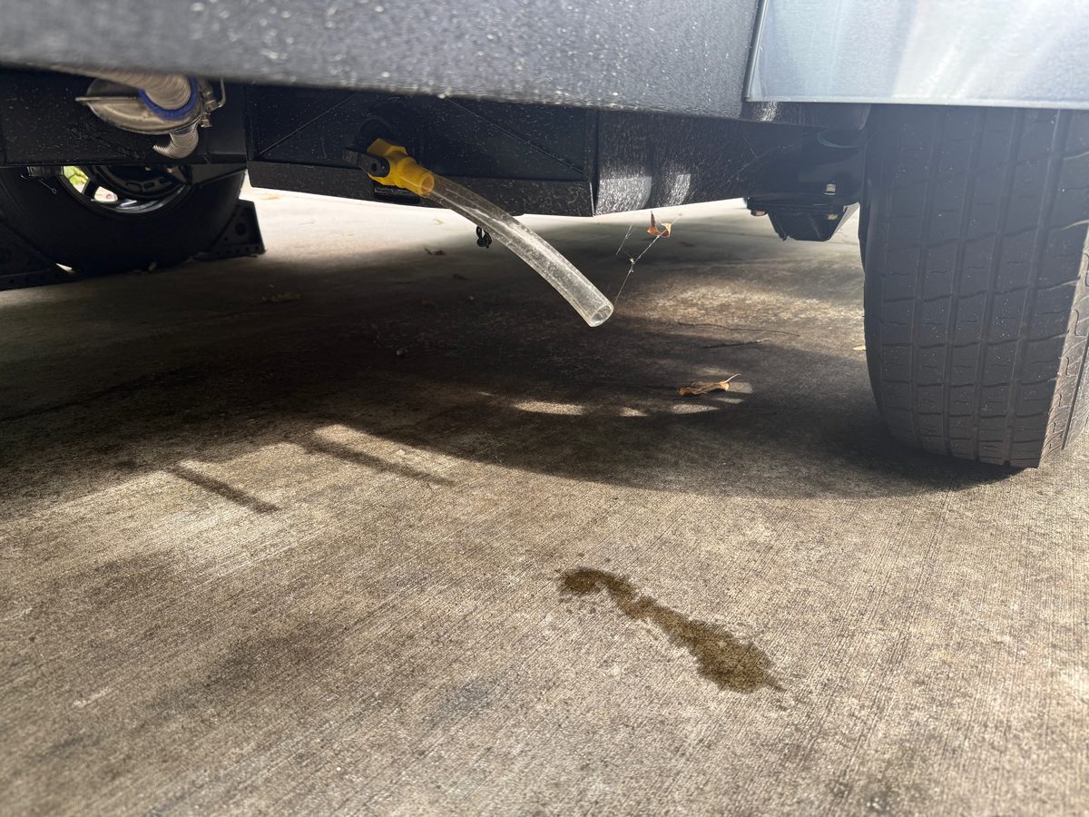 The underside of a teardrop trailer with a yellow valve open on a tank fitting and a clear hose draining onto a concrete driveway beside the tire