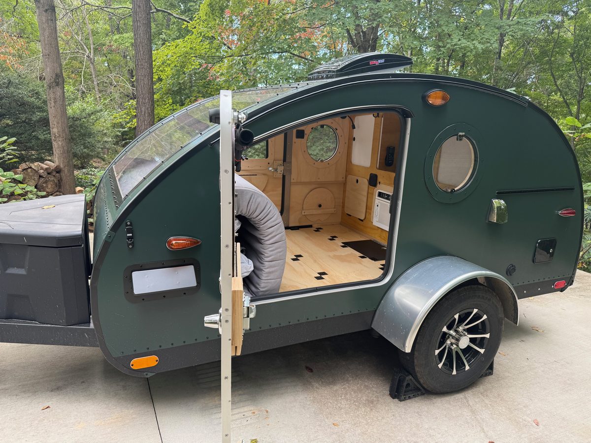 A green teardrop trailer parked on a driveway with its side door open and the birch cabin emptied out, a rolled gray bundle in the doorway and the wheel chocked
