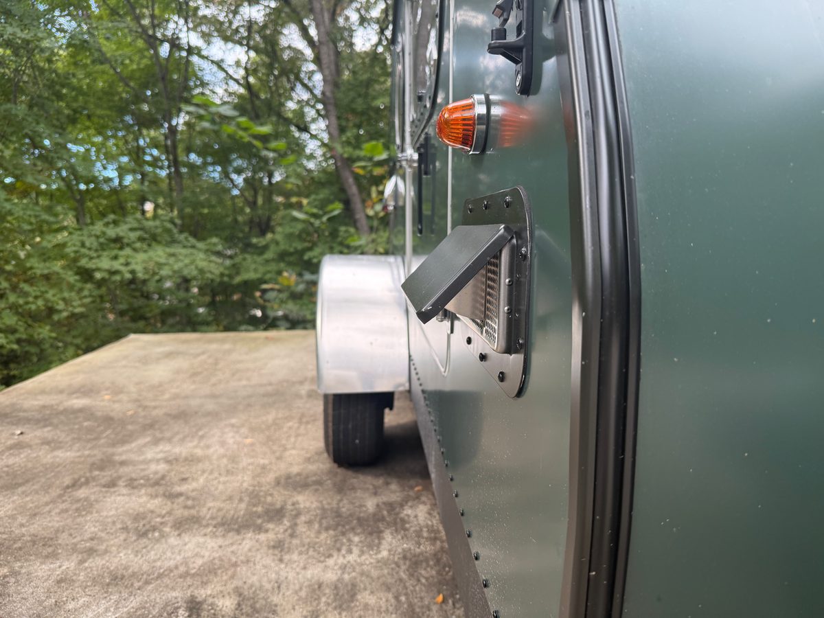 A small hinged vent flap propped open over a louvered grille low on the side of a green teardrop trailer